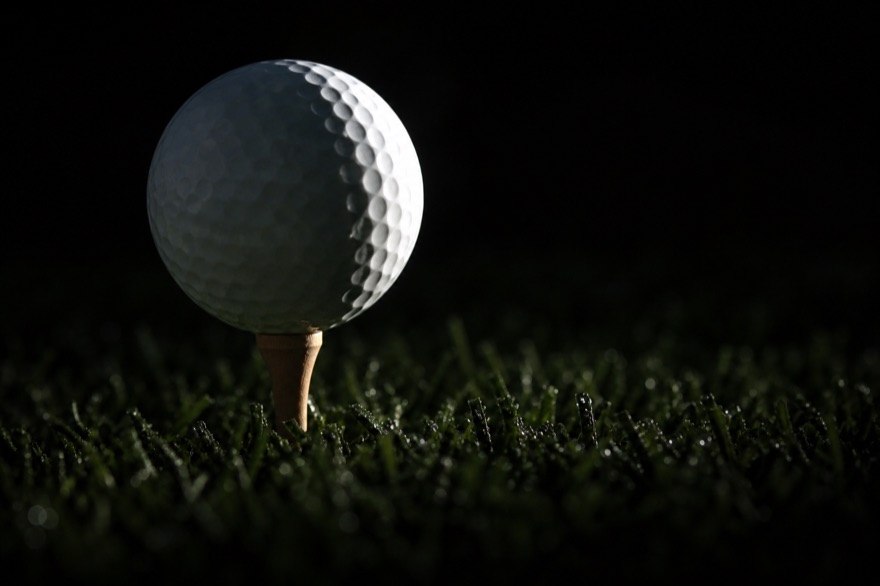 Golfball auf einem Holztee im taunassen Gras, von einem flachen Sonnenstrahl gestreift